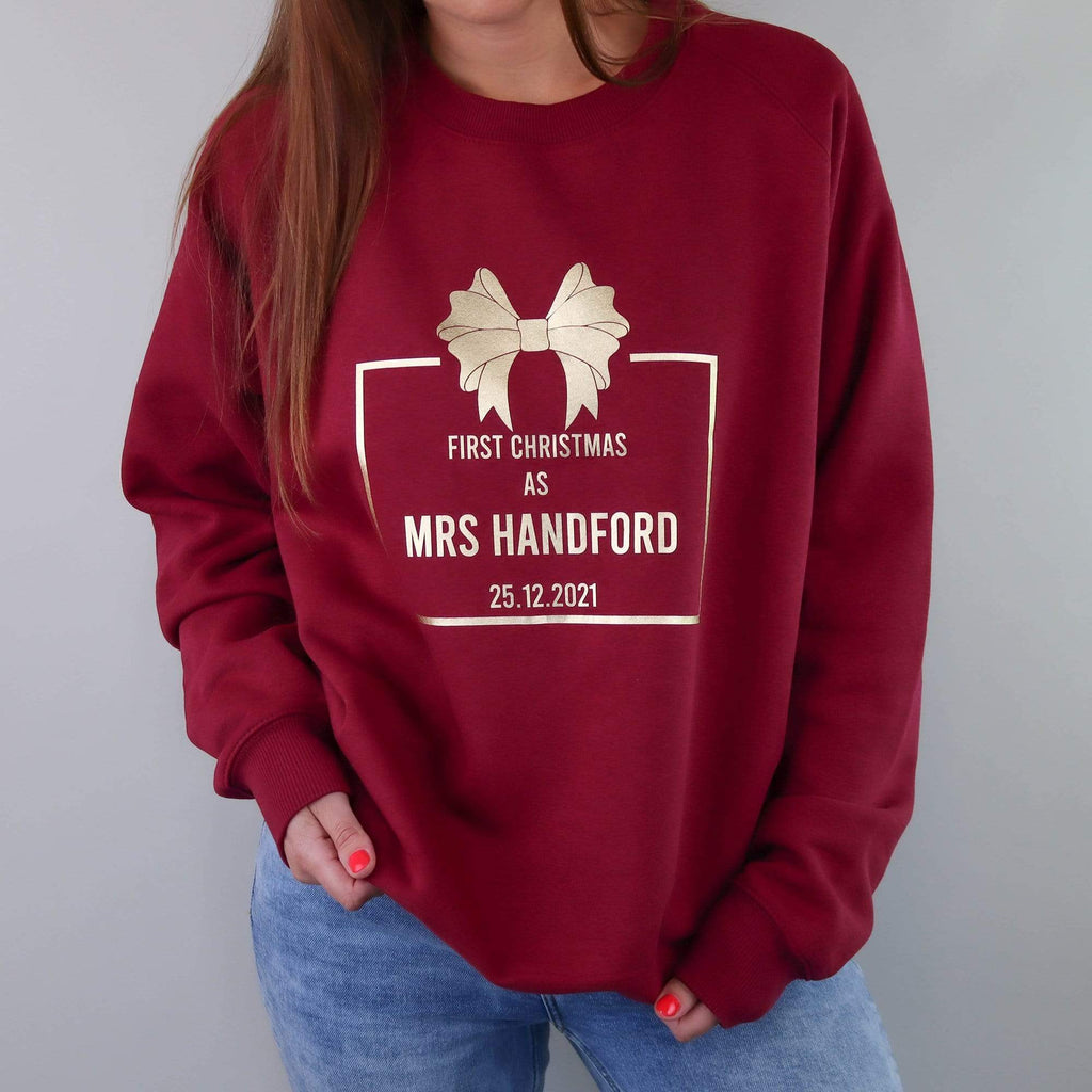 Woman wearing a burgundy sweater with gold text reading 'First Christmas as Mrs' and personalised with name and date. Also wearing blue denim jeans. By Original Monkey Gifts.