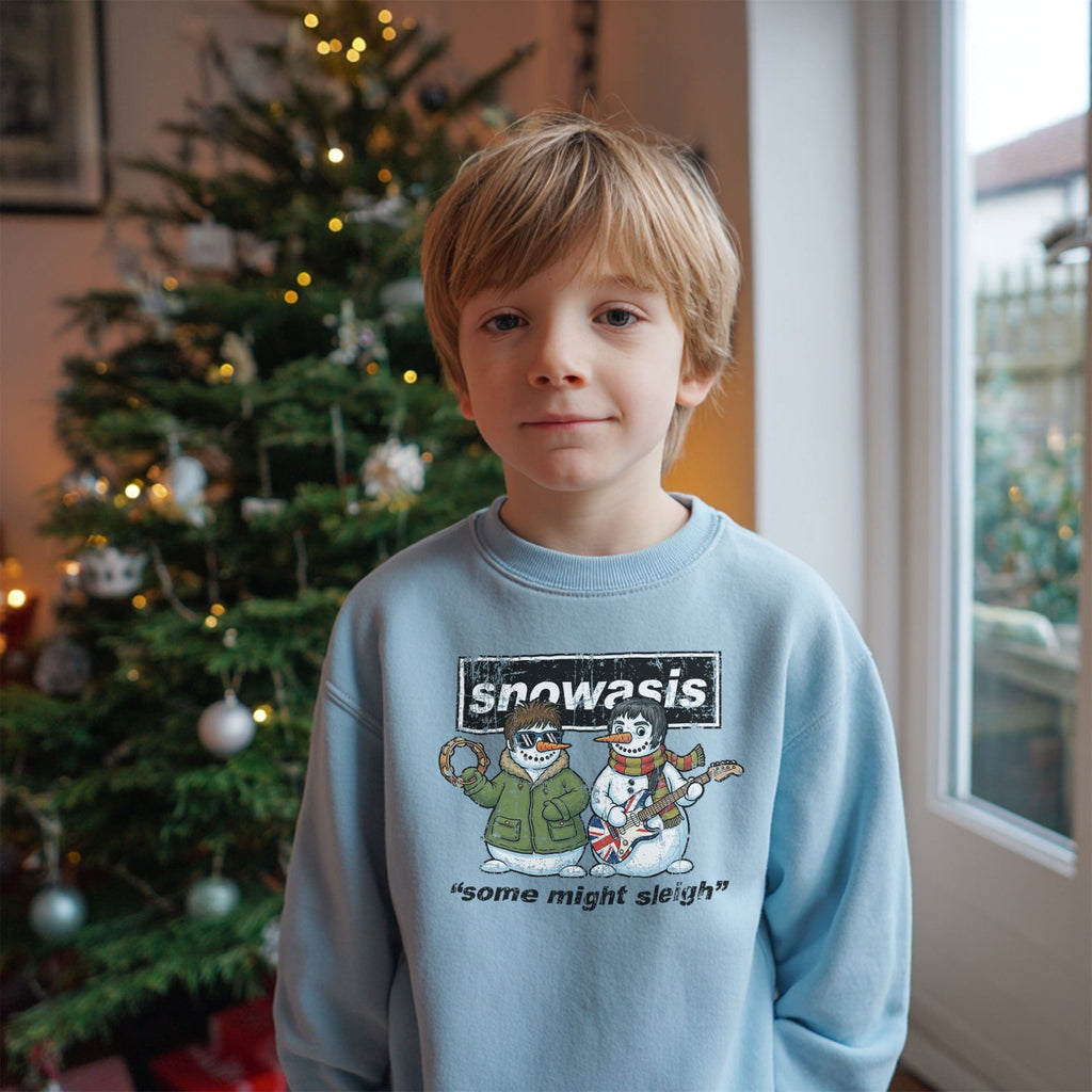 Young boy standing in front of a decorated Christmas tree, wearing a light blue sweatshirt with a festive design featuring two snowmen styled as a parody of the band Oasis. The text on the sweatshirt reads “Snowasis – some might sleigh.”