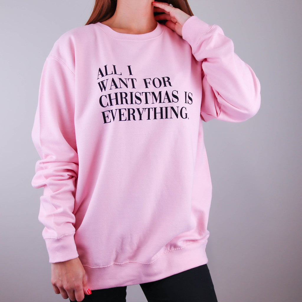 Woman wearing a pink Christmas jumper with text across the front that reads 'All I want for Christmas is everything' worn with black denim jeans. The message can be personalised and the jumper is made my Original Monkey Gifts.