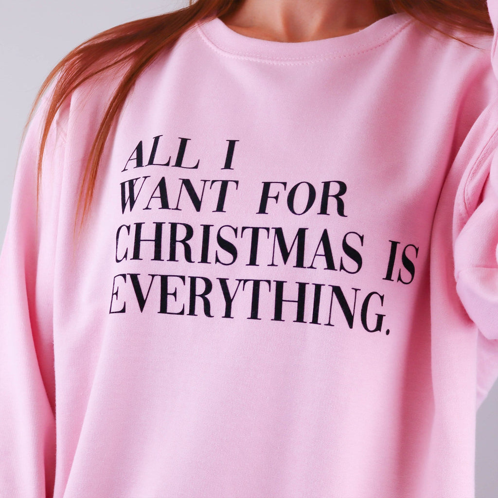 Woman wearing a pink Christmas jumper with text across the front that reads 'All I want for Christmas is everything' worn with black denim jeans. The message can be personalised and the jumper is made my Original Monkey Gifts.
