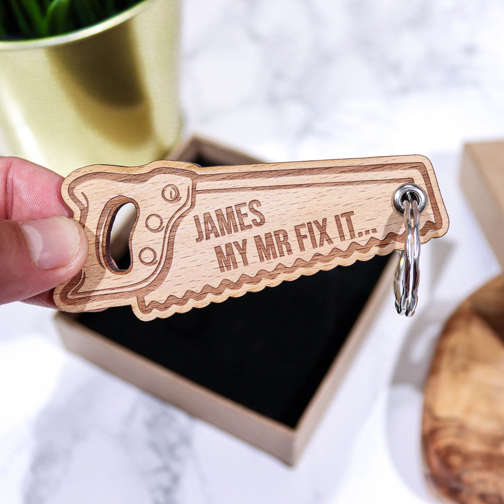 Man holding a saw shaped keyring that has a personalised message that reads 'James my mr mix it' by Original Monkey Gifts. A gift box and worktop are also pictured.
