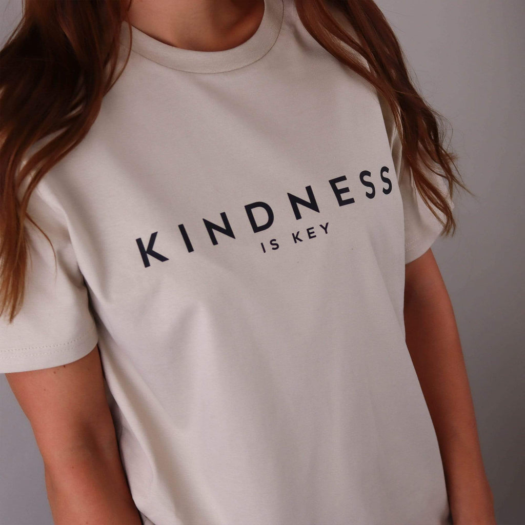 Woman wearing a nude t shirt with text that reads 'kindness is key' in black by Original Monkey Gifts. Woman also wears blue denim jeans, silver watch and white gold engagement and wedding rings.