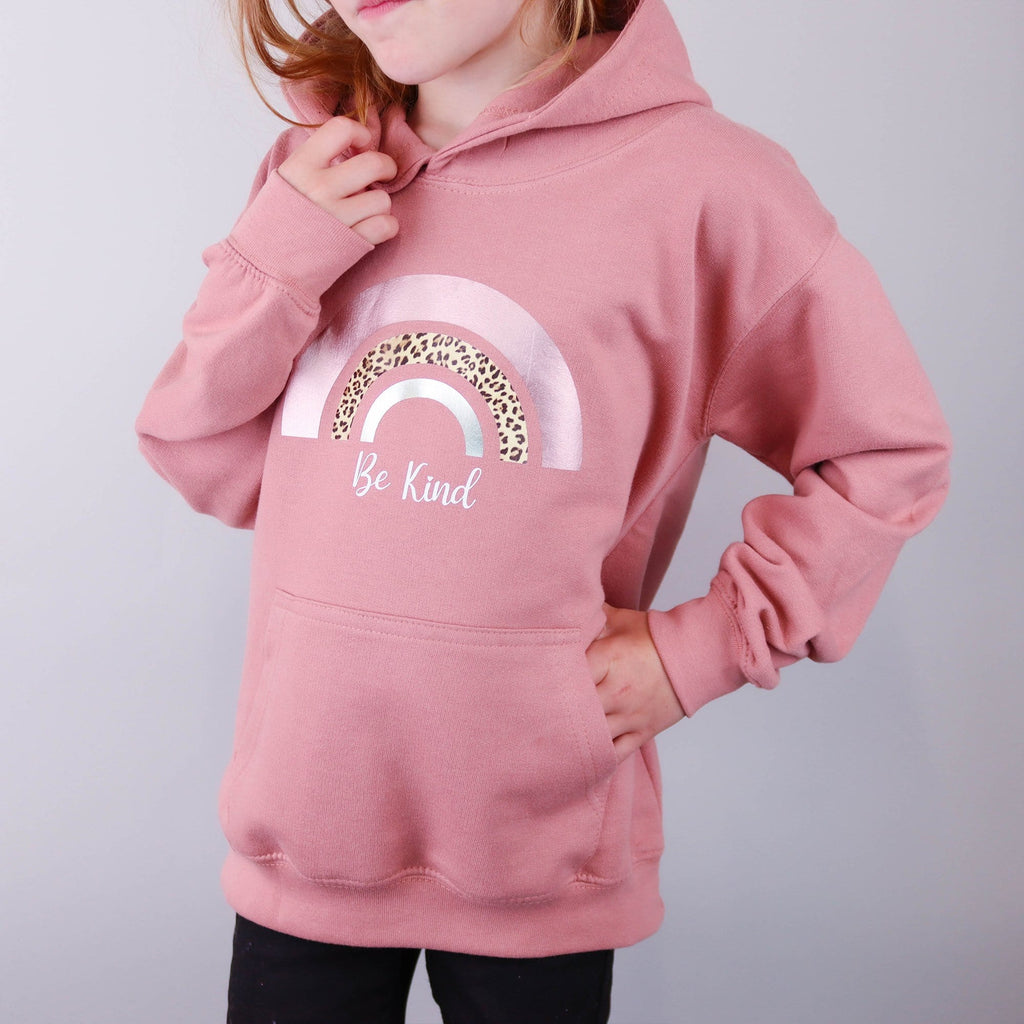 Child wearing a dusty pink hoodie with a rainbow print and saying be kind