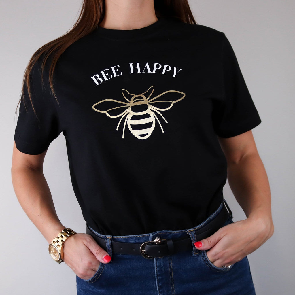 Woman wearing a black t shirt with a gold bee design and white text in a curve that reads 'bee happy' by Original Monkey Gifts. Woman also wears blue denim jeans, black leather belt and gold watch.