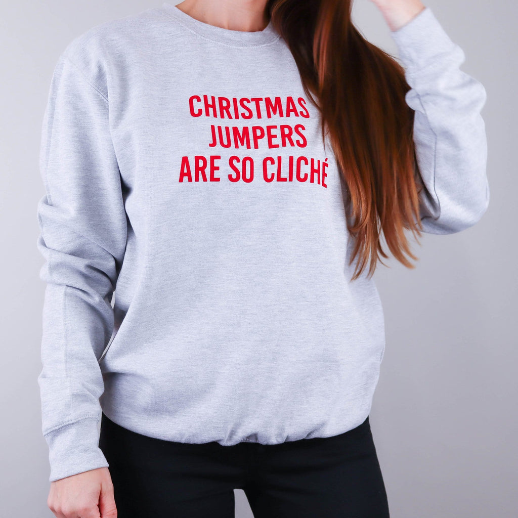 Woman wearing a grey sweater with red text that reads 'Christmas jumpers are so cliche' by Original Monkey Gifts. Woman also wears black denim jeans.