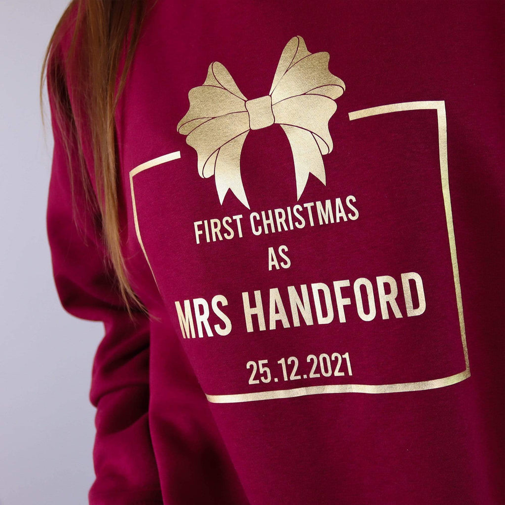 Woman wearing a burgundy sweater with gold text reading 'First Christmas as Mrs' and personalised with name and date. Also wearing blue denim jeans. By Original Monkey Gifts.