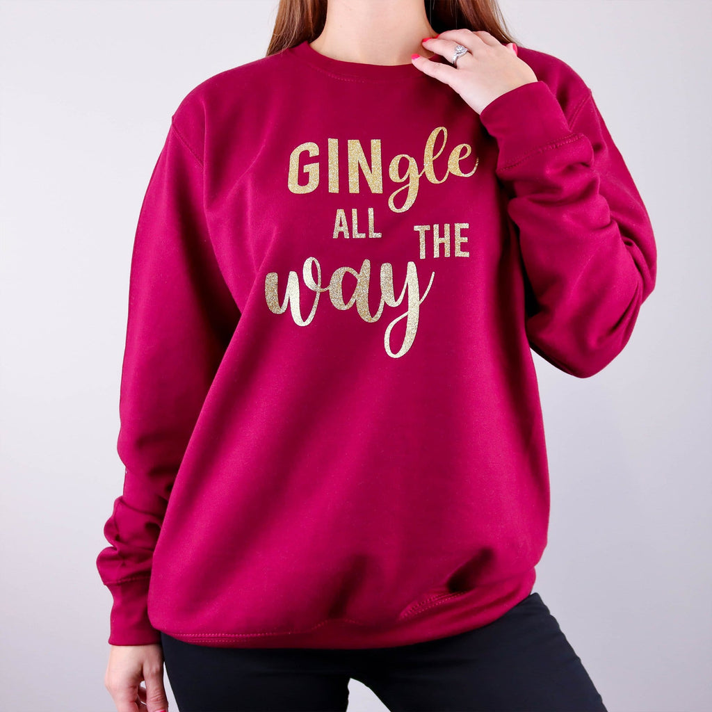 Woman wearing a burgundy Christmas jumper with text that reads 'GINgle all the way' in gold glitter by Original Monkey Designs. Woman also wears black denim jeans and white gold wedding and engagement rings.