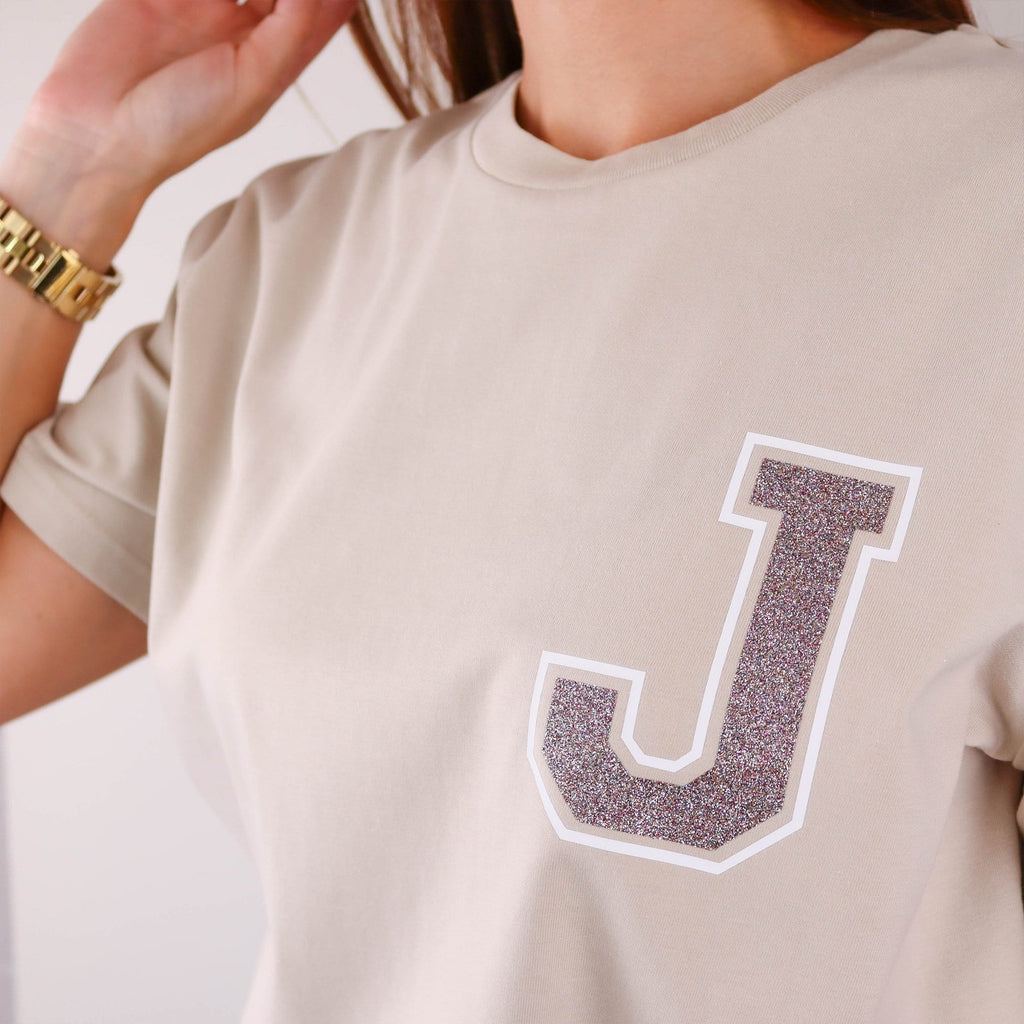 Woman wearing nude t shirt with a glittery initial 'J' on the left hand side by Original Monkey Gifts. Woman also wears blue denim jeans, brown leather belt and gold watch.