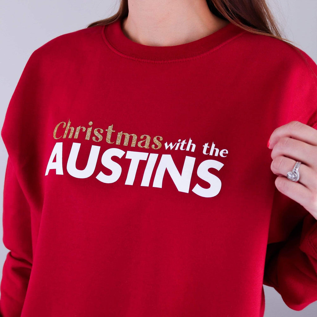 Woman wearing red jumper with text reading 'Christmas with the...' that can be personalised with any surname by Original Monkey. Woman also wearing black denim jeans and white gold wedding and engagement rings.