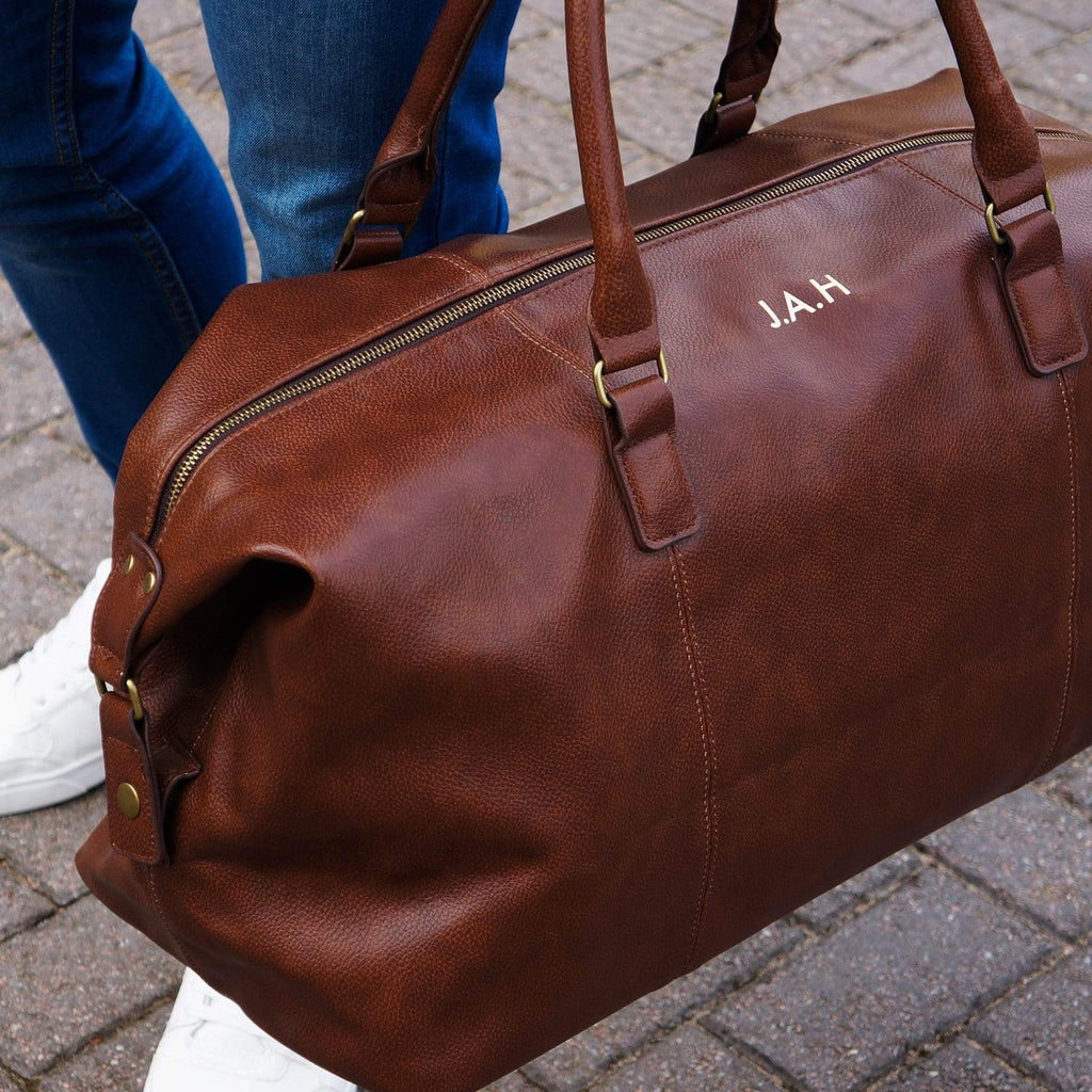Man carrying a brown PU leather bag with personalised initials in gold by Original Monkey Gifts. Man wears blue denim jeans and white trainers.