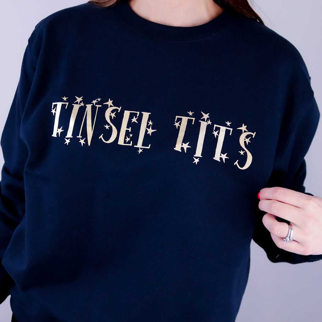 Woman wearing a navy sweater with gold writing that reads 'tinsel tits' made by Original Monkey Gifts.
