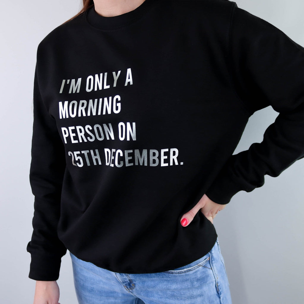 Woman wearing black sweater with silver text reading 'I'm only a morning person on 25th December' with blue denim jeans. By Original Monkey.